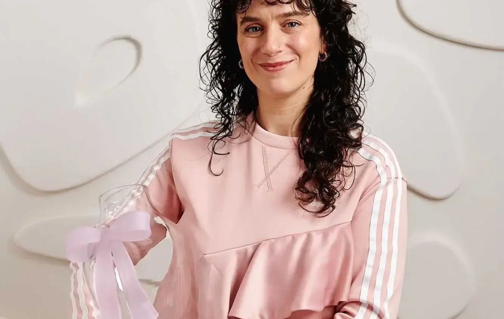 Eine Person mit lockigem dunklem Haar in einem rosafarbenen Outfit hält einen durchsichtigen Preis mit einer Schleife und steht vor einer weiß strukturierten Wand - ein inspirierender Moment im Linda Pro Concept Store.