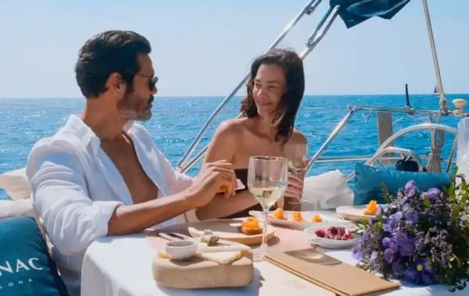Un hombre y una mujer sentados a la mesa en un barco, disfrutando de comida y bebida con el océano de fondo: una experiencia inolvidable en Suite en un día soleado.
