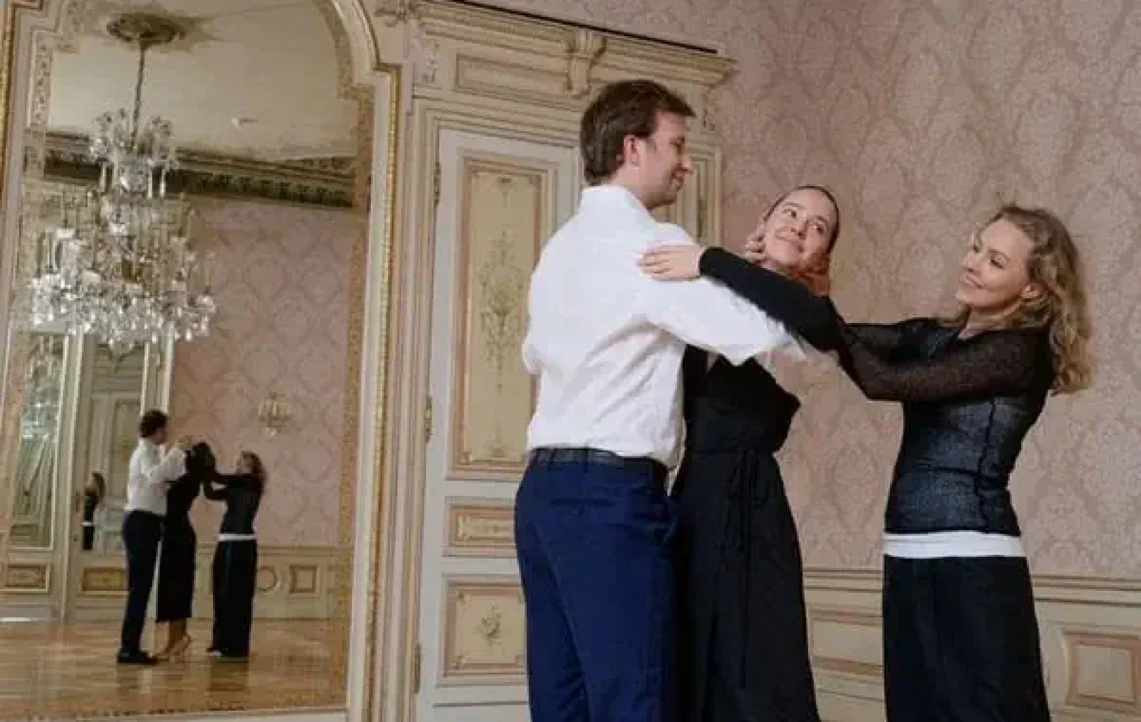 Three people practice ballroom dancing in an ornate room with a chandelier and large mirror, preparing for a glamorous Night At The Ball.
