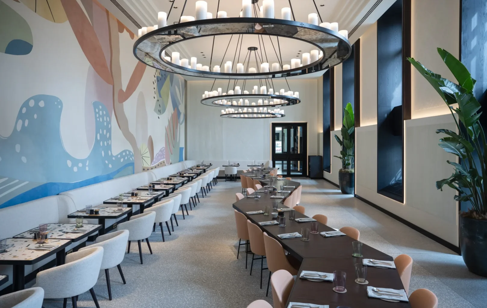 Modern restaurant interior at Almanac Palais Vienna with two long dining tables, light-colored chairs, large circular chandeliers, abstract wall art, tall plants, and floor-to-ceiling windows.