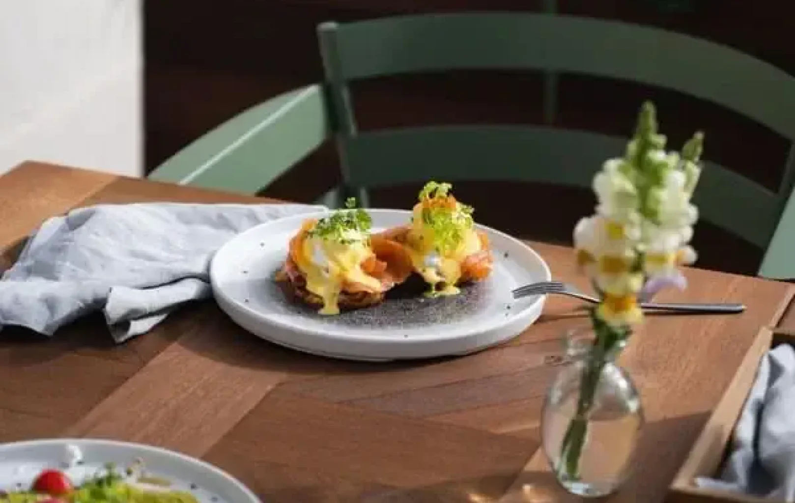 A plate of eggs Benedict on an English muffin sits on a wooden table, capturing the essence of Summer Elegance beside a vase with flowers and another plate of food.