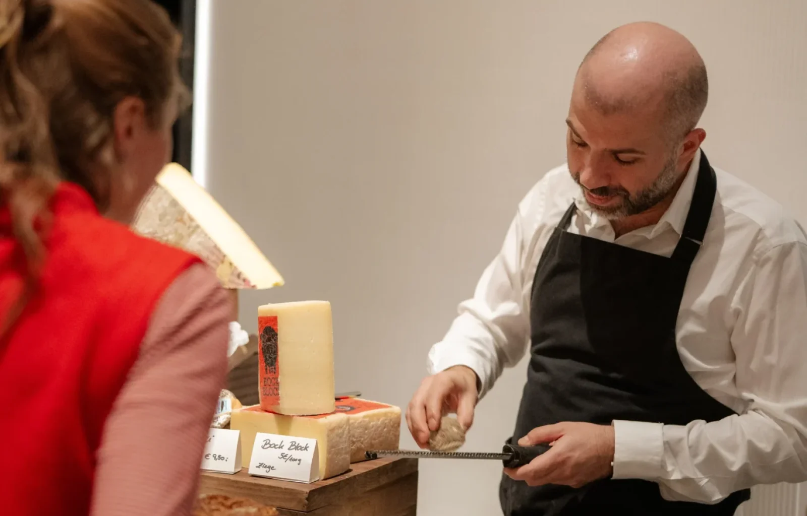 A man in an apron slices bread at a table with various types of bread and cheese, while a woman in a red jacket stands nearby, savoring flavors that evoke the charm of culinary destinations.