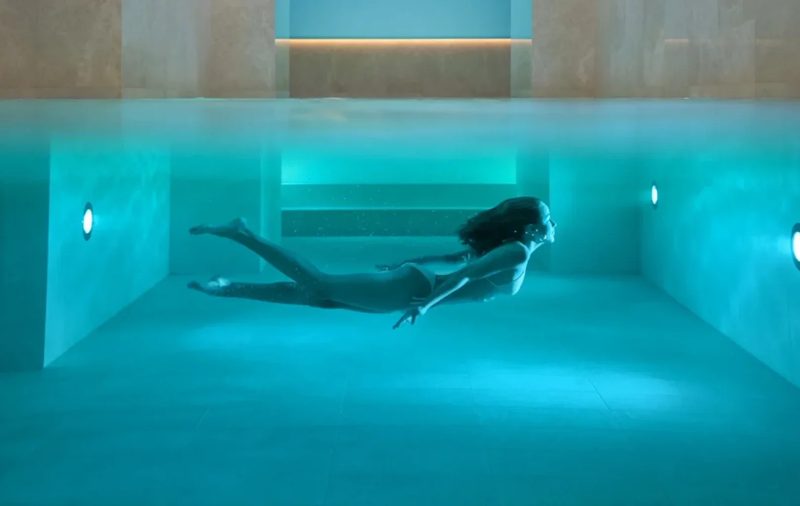 Woman swimming underwater in a modern indoor spa pool, illuminated by blue lights for a soothing wellness experience, with stairs visible on the left side.