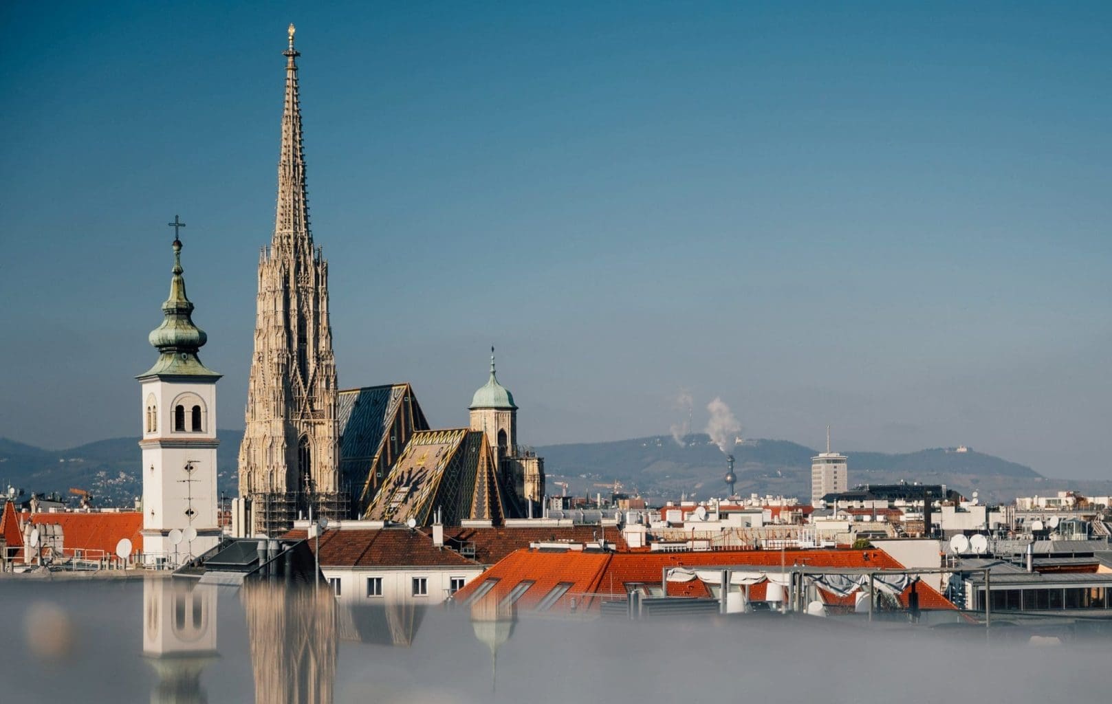 Panoramic view of Vienna’s cityscape from almanac palais vienna hotel featuring St. Stephen’s Cathedral with its tall spire and patterned roof under a clear blue sky.