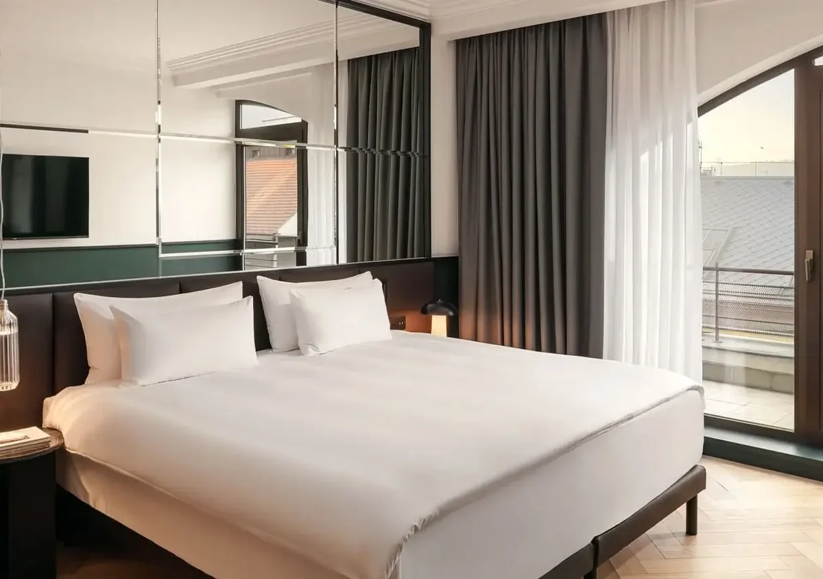 Experience our Executive Suite: a modern hotel room featuring a large bed with white linens, mirrored wall, wall-mounted TV, bedside tables, and glass doors opening to a private balcony.