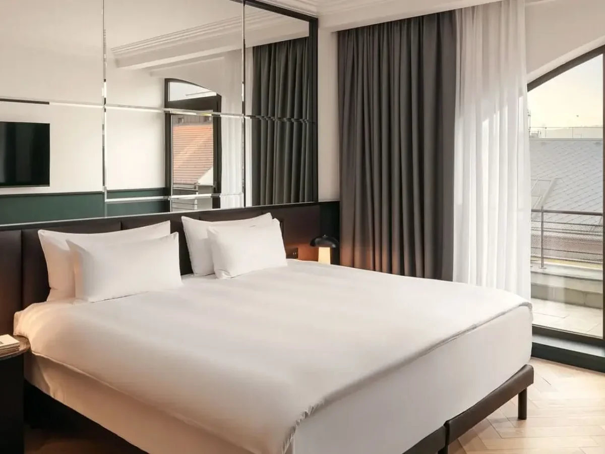 Experience our Executive Suite: a modern hotel room featuring a large bed with white linens, mirrored wall, wall-mounted TV, bedside tables, and glass doors opening to a private balcony.