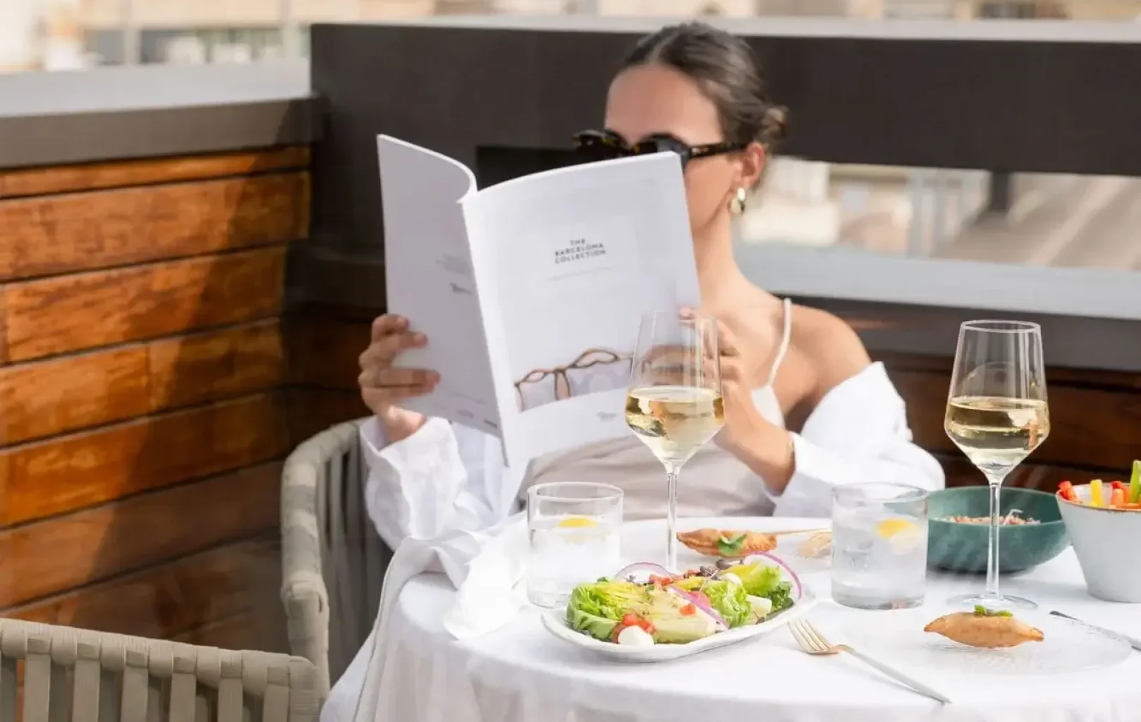 Una mujer disfruta de una Suite Experience en la mesa de un restaurante al aire libre con ensaladas, vino y agua, leyendo un menú mientras al fondo se ven edificios de la ciudad.