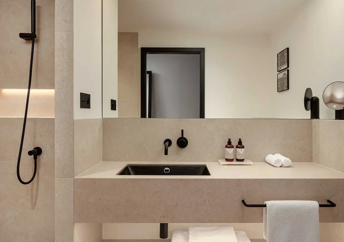 Modern bathroom with beige tiles, black fixtures, a wall-mounted sink, rolled towels, two soap dispensers, a large mirror, and a walk-in shower with a handheld showerhead.