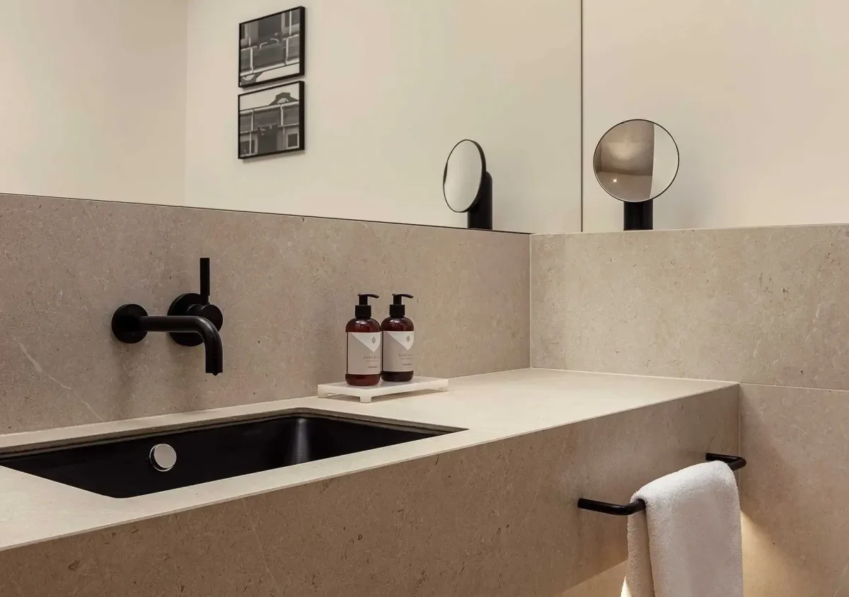 Modern bathroom with beige stone countertop, black sink and faucet, two soap dispensers, a towel hanging, and a round mirror on the counter. Two framed photos hang on the wall.