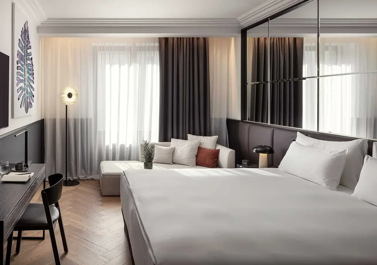 Modern hotel room with a large bed, desk and chair, sofa with cushions, floor lamp, and curtains covering tall windows.