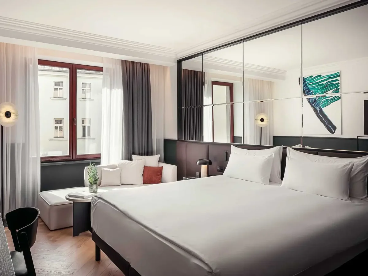 Modern Almanac X hotel room with a large bed, mirrored wall, sofa, two windows with sheer curtains, abstract artwork above the bed, and access to a private balcony.