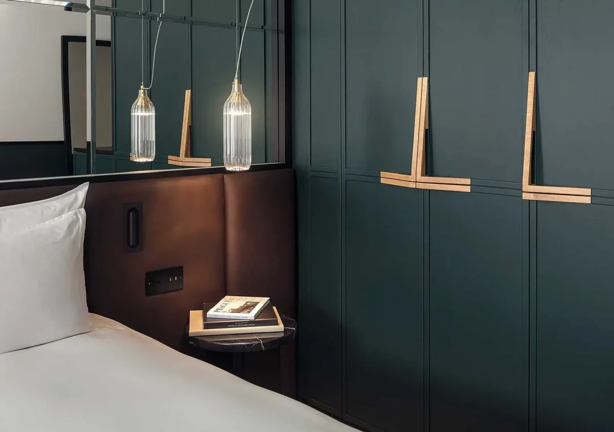 A modern hotel room with a white bed, a dark headboard, a small bedside table holding two books, pendant lights, and green paneled wardrobe doors with wooden handles.