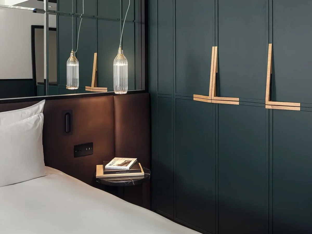 A modern hotel room with a white bed, a dark headboard, a small bedside table holding two books, pendant lights, and green paneled wardrobe doors with wooden handles.