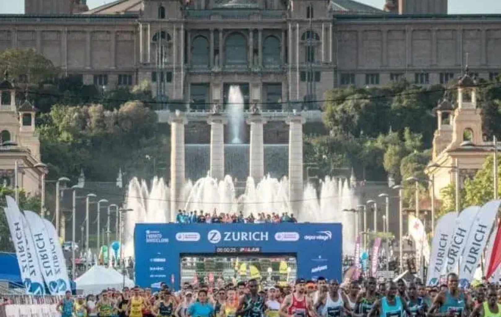 Una gran multitud de corredors de marató comença una cursa prop d'un edifici històric amb fonts, sota una pancarta blava de Zuric en un dia assolellat d'abril.
