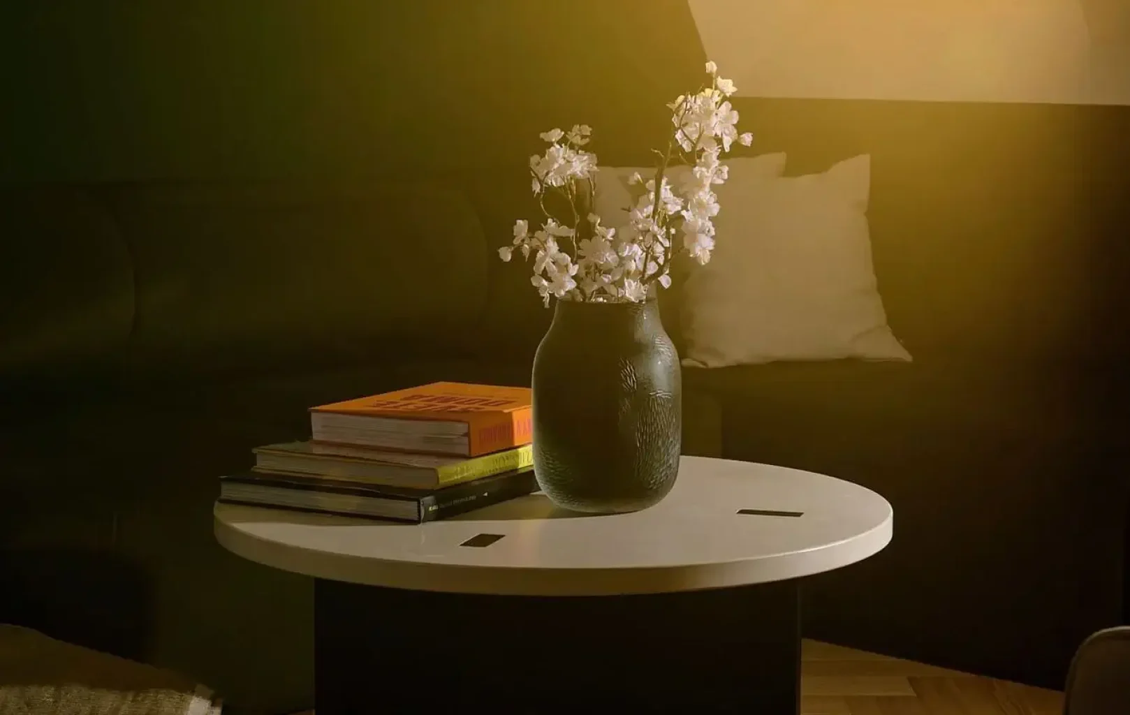 V prosluněném pokoji s tmavou pohovkou a polštářem, evokujícím eleganci prezidentského apartmá, stojí kulatý konferenční stolek s knihami a vázou Rücklův Křišťálový sen.