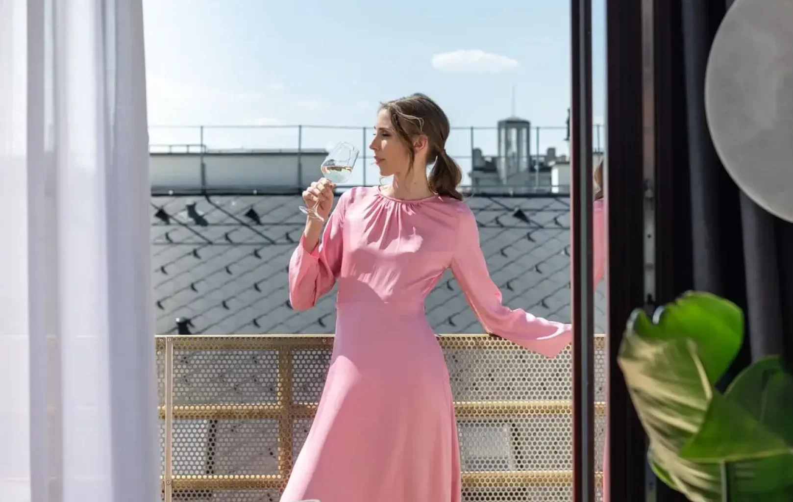 Eine Frau in einem langen rosa Kleid steht barfuß auf einem Balkon, hält ein Weinglas in der Hand und schaut an einem sonnigen Tag nach draußen.