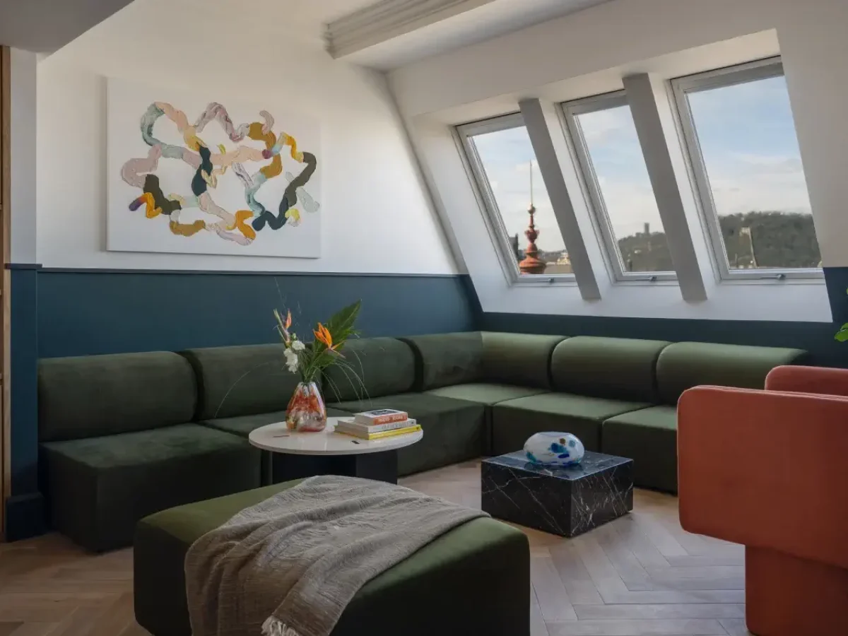 Modernes Wohnzimmer mit grünem Sektionssofa, orangefarbenem Sessel, abstrakter Wandmalerei und großen Fenstern, die an die Prager Hotels Almanac und Alcron erinnern, sowie einem Marmorcouchtisch und einem kleinen runden Tisch mit Blumen.