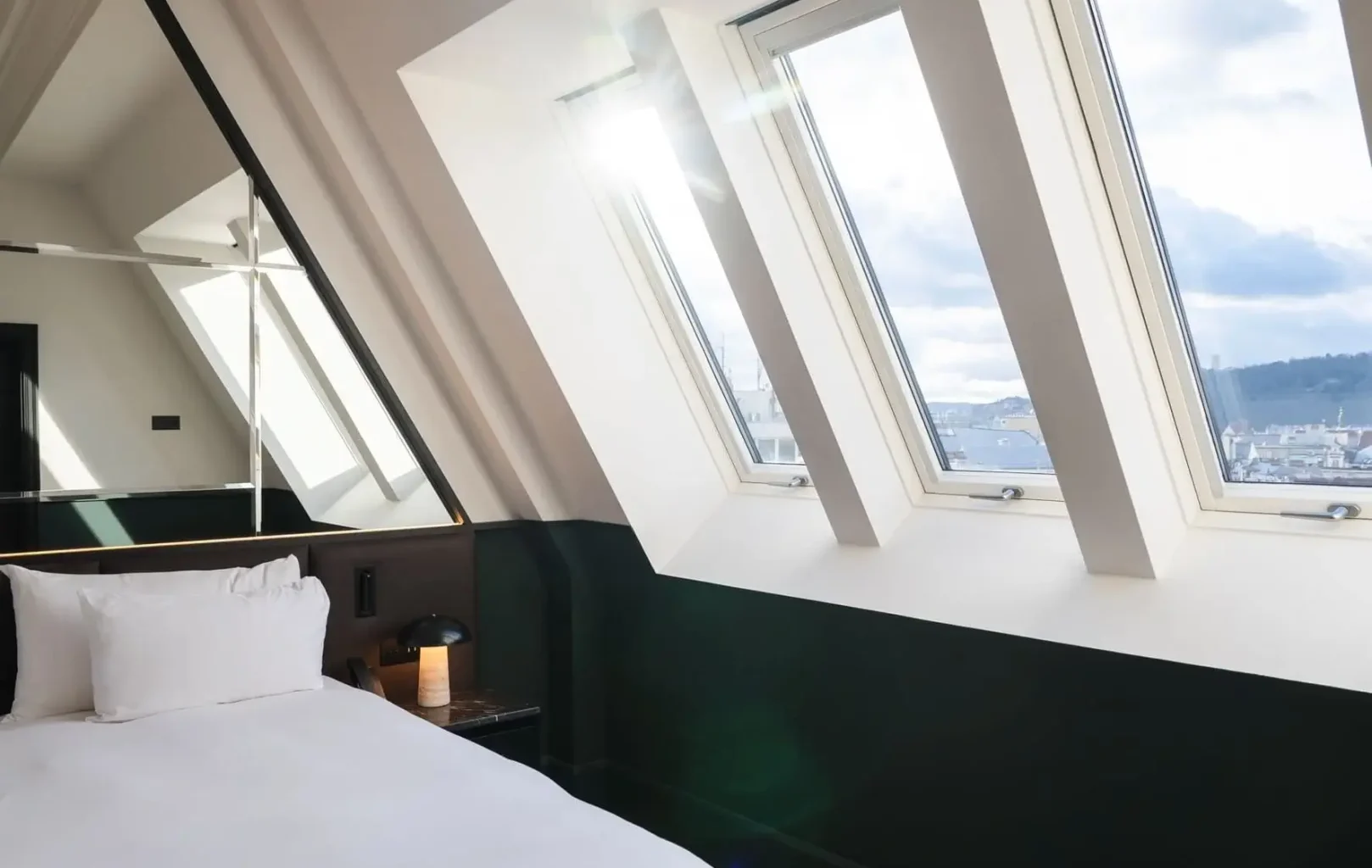 A neatly made bed beside large slanted windows with sunlight streaming in, overlooking a cityscape, in the modern Castle View Suite at Almanac X.
