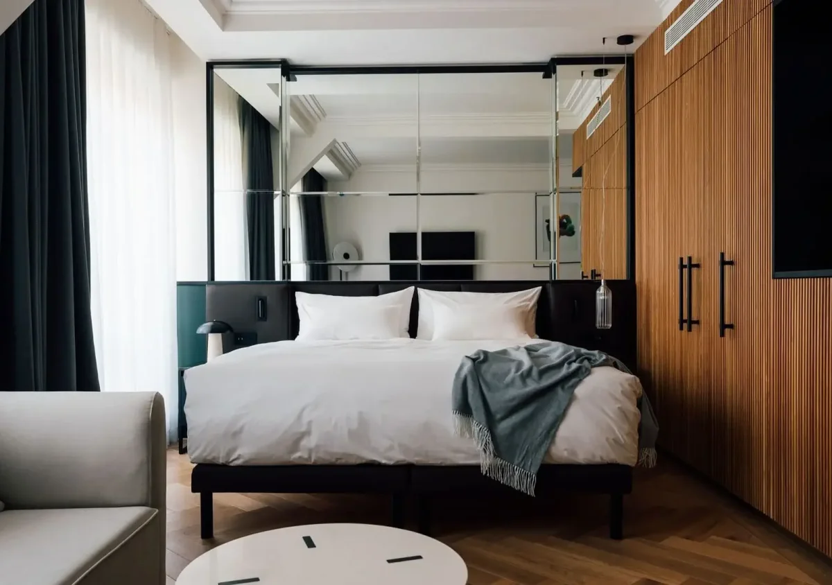 Modern Junior Suite featuring a neatly made double bed, gray throw blanket, wood-paneled wardrobes, large mirror, sofa, and round white table. Natural light flows in through a sheer curtain for a bright and inviting atmosphere.