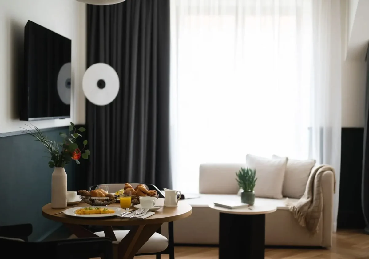 A modern Junior Suite living room features a round table set for breakfast, a white sofa, a coffee table with a plant, and large windows with sheer curtains.