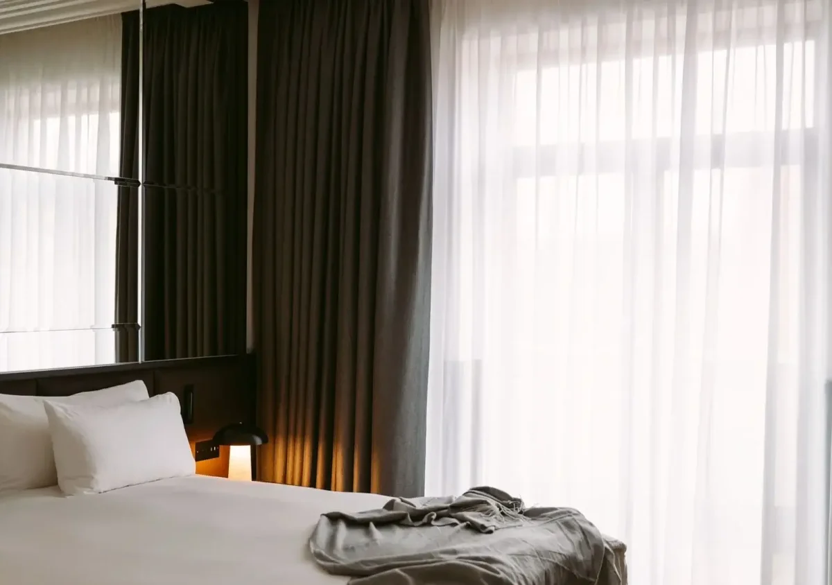 A neatly made bed with white linens and pillows sits beside a window with sheer and blackout curtains in this Executive Suite, allowing natural light into the room; a blanket is draped at the foot of the bed.