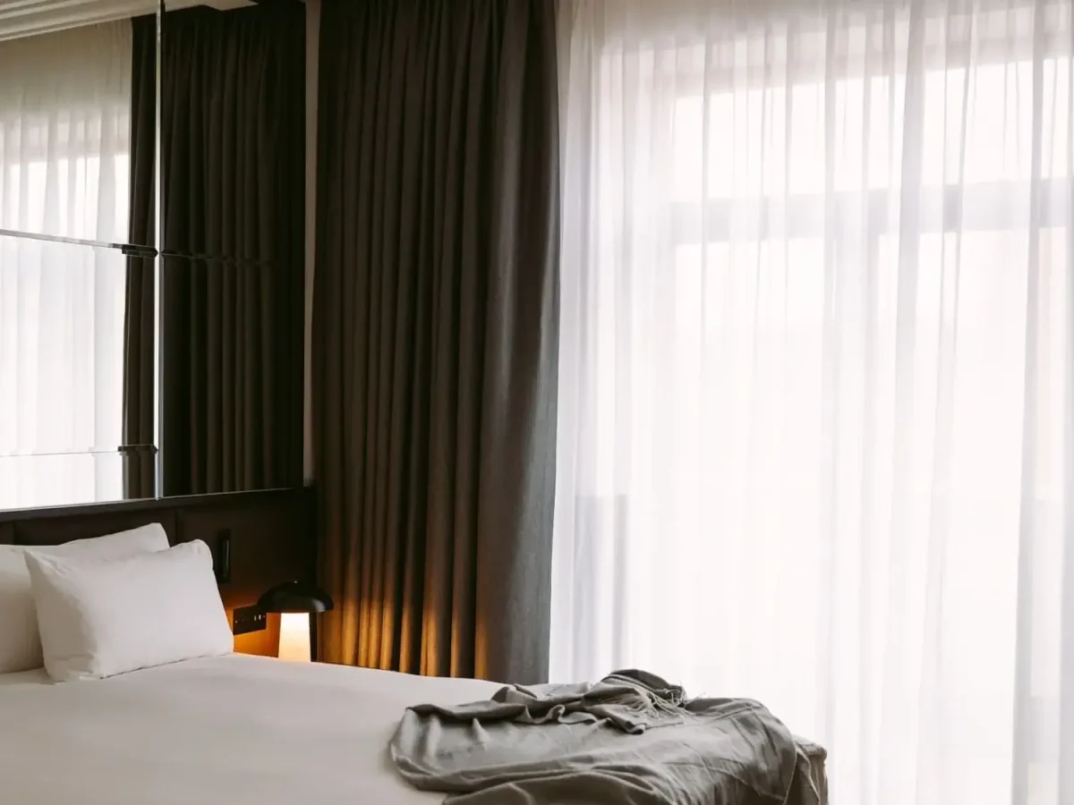 Úhledně ustlaná postel s bílým povlečením a polštáři leží vedle okna s průsvitnými závěsy a zatemňovacími záclonami v měkce osvětlené moderní ložnici Apartmá Executive.