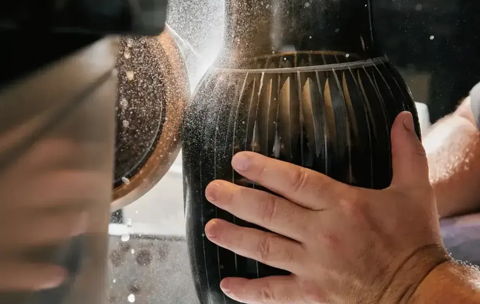 A person’s hand holds a black ceramic vase while a spinning tool cuts vertical grooves into its surface, creating fine dust—a captivating moment often witnessed during Signature Tours of local artisan workshops.