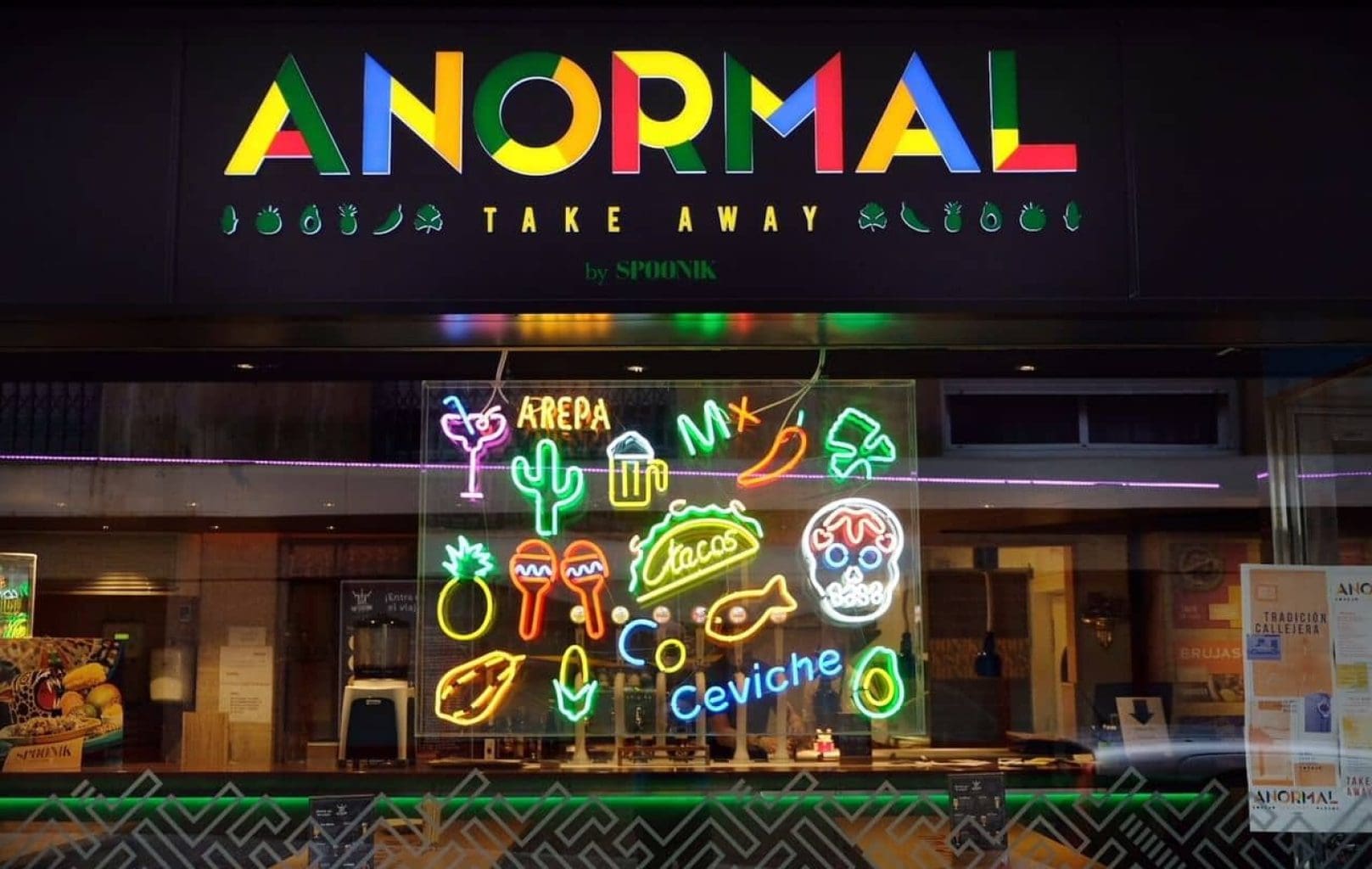 Storefront with an "ANORMAL TAKE AWAY" sign and neon window display featuring tacos, cactus, skulls, chili peppers, and words like “Arepa” and “Ceviche”—a vibrant showcase of Anormal Food.