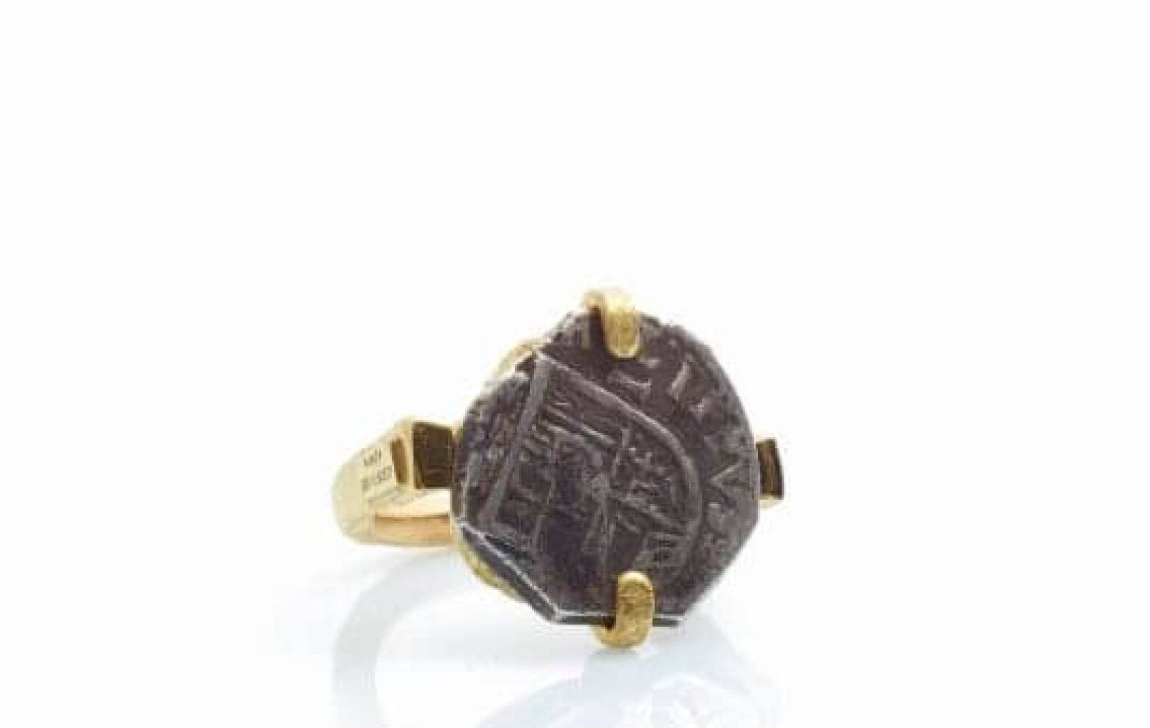 A gold ring by Assumpta Bou featuring a dark, irregularly shaped coin or artifact as its centerpiece, elegantly held by four prongs.