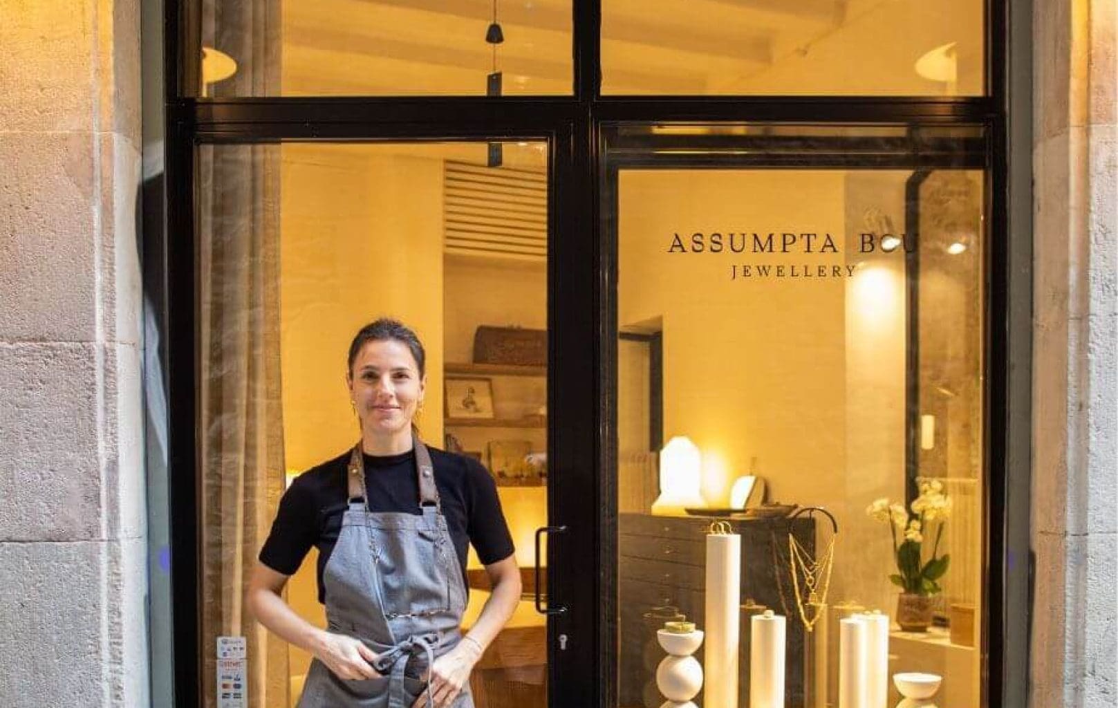 A person wearing an apron stands in front of a jewelry shop with glass windows displaying decorative items and the sign "ASSUMPTA B JEWELLERY" by Assumpta Bou visible behind them.