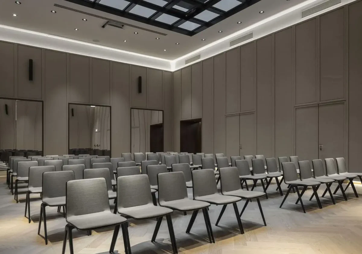 Ein moderner Konferenzraum für Meetings und Veranstaltungen, mit leeren grauen Stuhlreihen an der Stirnseite, hohen Decken, neutralen Wänden und einem großen Oberlicht über dem Kopf.