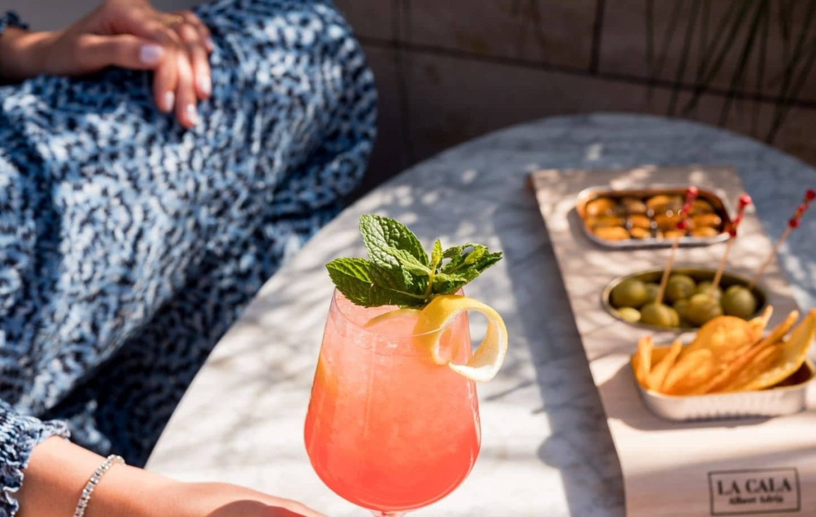 A person holds a pink cocktail garnished with mint and lemon at a marble table at Azimuth Rooftop Bar, surrounded by bowls of olives, chips, and nuts in sunlight.