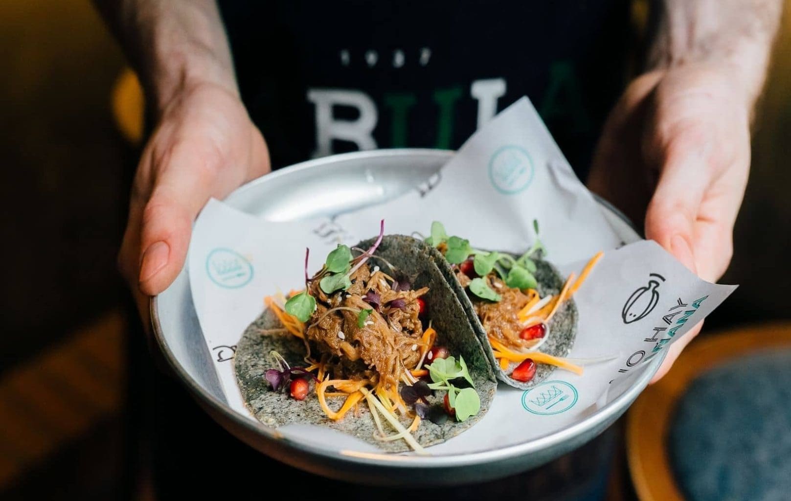 Una persona sosté un plat amb dos tacos farcits de carn esmicolada, microverdures i verdures, servits en paper imprès al BABULA BAR 1937.