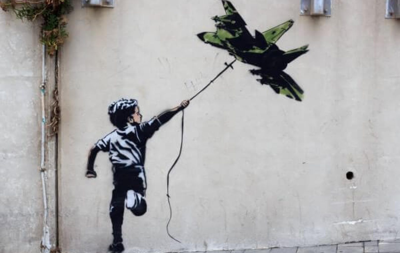 Un mural al estilo de Banksy muestra a un niño sujetando una cuerda atada a un globo con forma de avión de combate, pintado sobre una pared lisa: un evocador ejemplo de arte de la protesta.