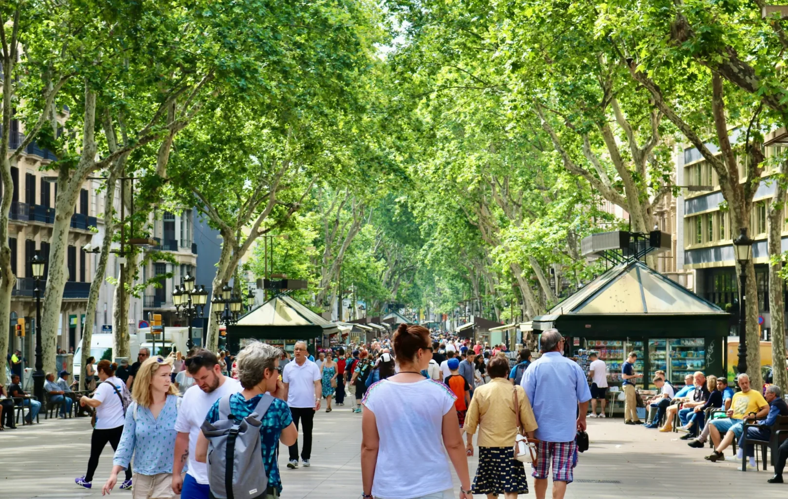 A busy pedestrian street lined with trees and kiosks near L’Auditori in Barcelona, with people walking and socializing on a sunny day.