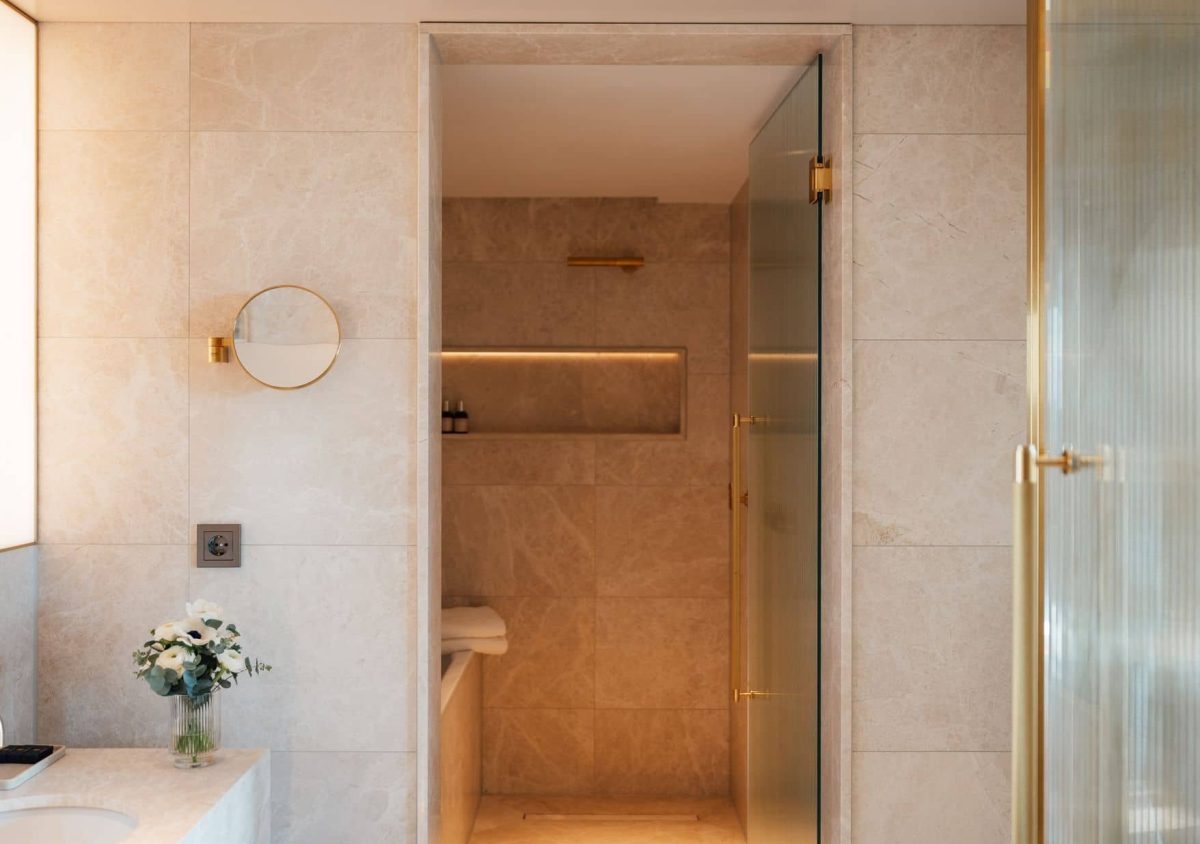 Modern Almanac Zimmer Badezimmer mit beigen Fliesen, einer Glastür in der Dusche, goldenen Armaturen, einem runden Wandspiegel und einem kleinen Blumenstrauß auf dem Waschbecken.