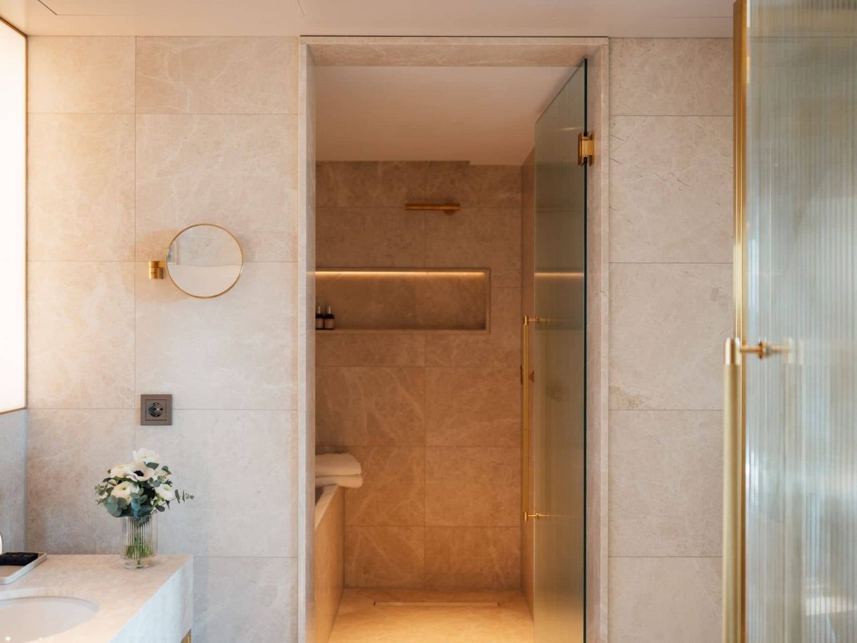 Modern Almanac Zimmer Badezimmer mit beigen Fliesen, einer Glastür in der Dusche, goldenen Armaturen, einem runden Wandspiegel und einem kleinen Blumenstrauß auf dem Waschbecken.