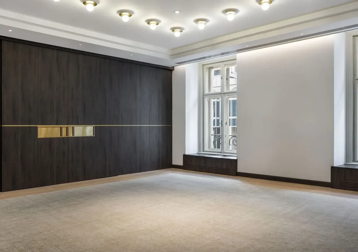 Ein geräumiger, leerer Raum mit großen Fenstern, einem hellen Teppich, dunkler Holzvertäfelung an einer Wand und mehreren Deckenleuchten.