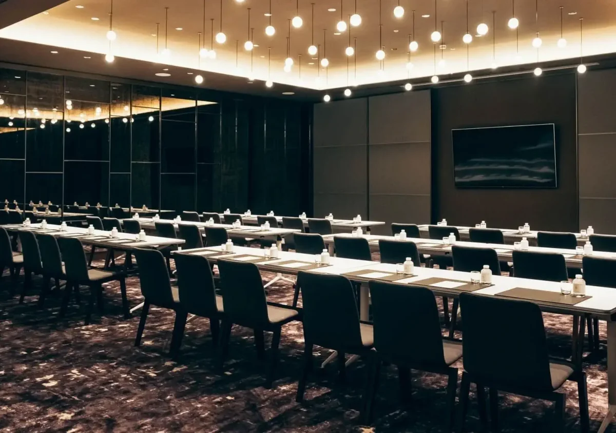 Moderner Konferenzraum mit Tisch- und Stuhlreihen, Notizblöcken und Wasserflaschen auf jedem Tisch, der unter Deckenbeleuchtung und einem großen Wandbildschirm den perfekten Raum für Vorveranstaltungen bietet.