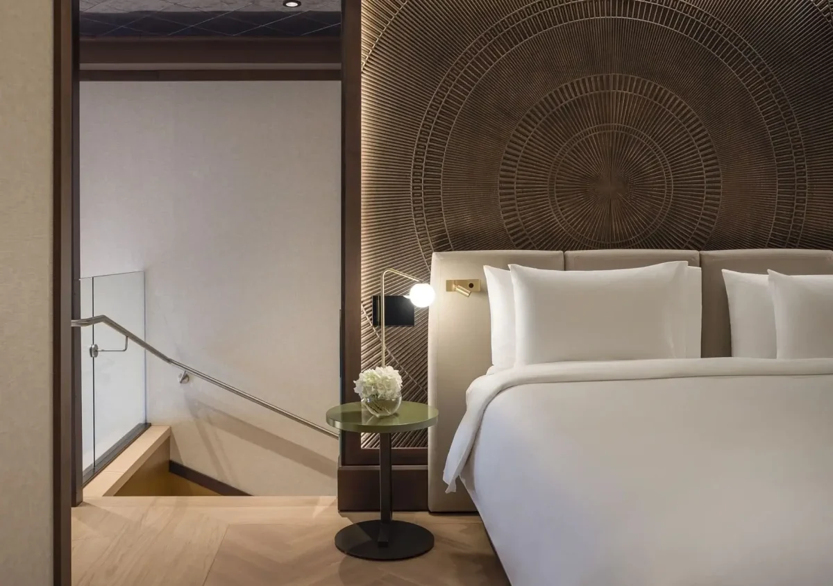 A modern Duplex Suite featuring a white bed, circular patterned headboard, small round side table with a flower arrangement, and stairs with a glass railing leading down.