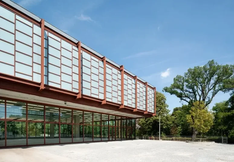 Modern glass and steel building with large windows and a flat roof, featuring elegant rooms and suites, set beside trees on a clear day.
