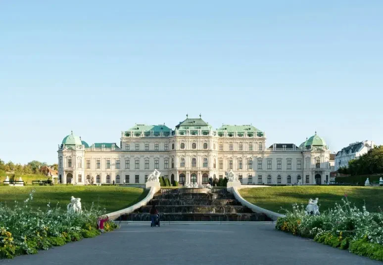A grand baroque palace with green domes, cascading fountains, and landscaped gardens under a clear blue sky.