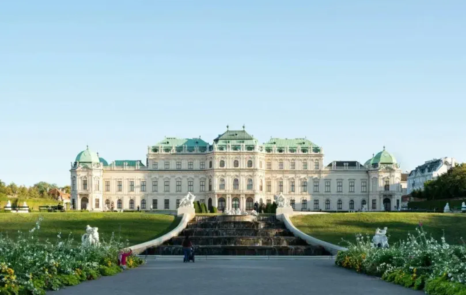 A grand baroque palace with green domes, cascading fountains, and landscaped gardens under a clear blue sky.