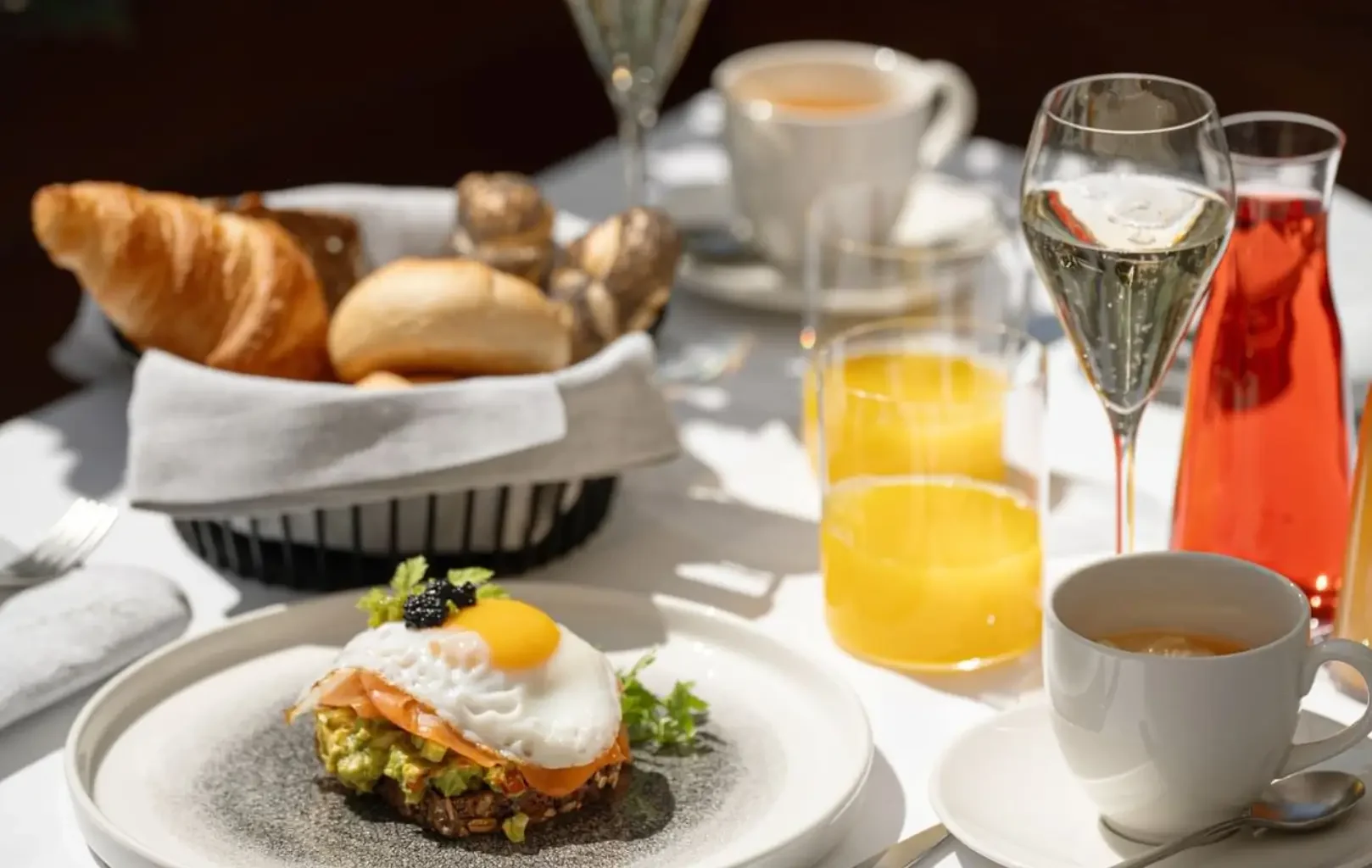 A breakfast table awaits with croissants, bread rolls, orange juice, coffee, a red drink, champagne, and a plate of avocado toast topped with salmon, a fried egg, and caviar. Start your day right—Breakfast With Us.