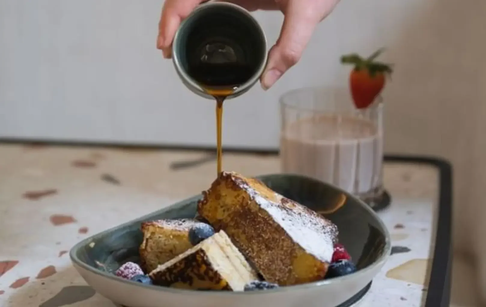 A hand pours syrup over slices of French toast topped with powdered sugar and berries; a glass of chocolate milk with a strawberry completes this inviting breakfast scene. Start your day right—enjoy Breakfast With Us.