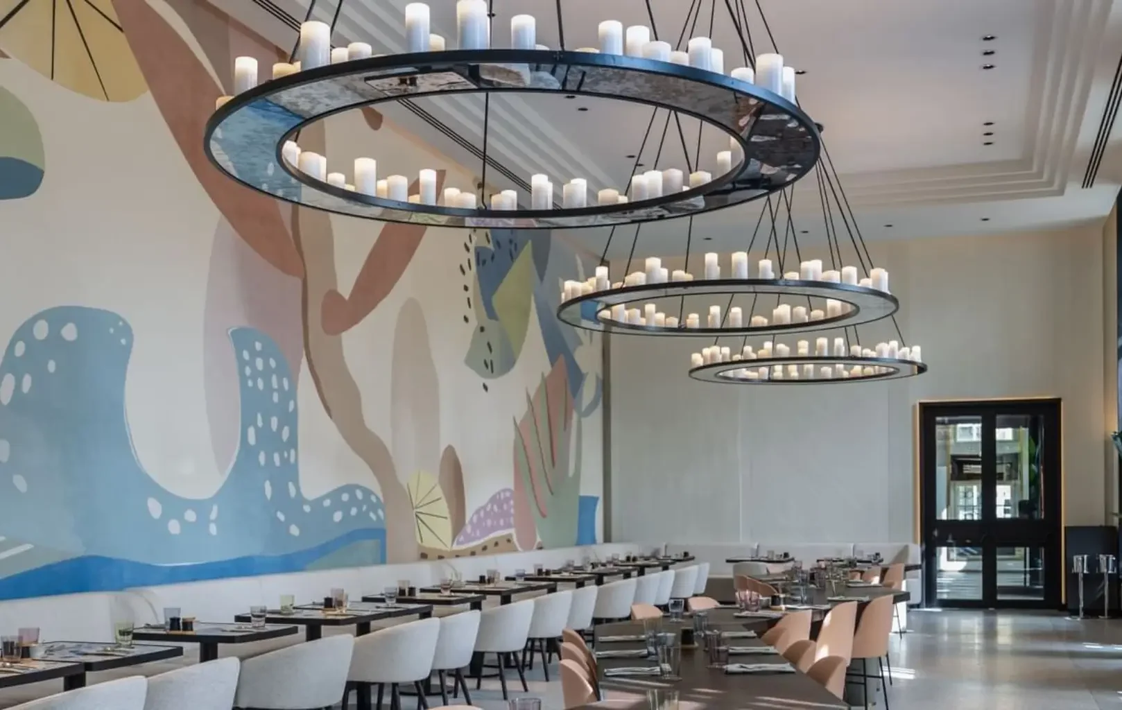 Modern dining area with long tables, cushioned chairs, large circular chandeliers with candles, and a colorful abstract mural on the wall—perfect for starting your day. Enjoy Breakfast With Us in this inviting space.