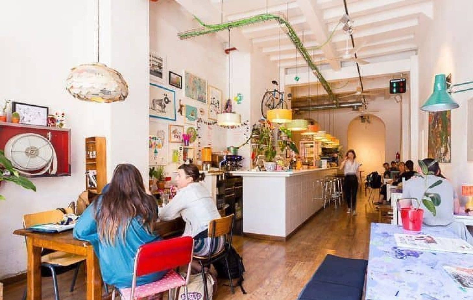 Bright, cozy café with eclectic décor, artwork on white walls, plants, and customers seated at tables and the counter. Natural light fills the space.
