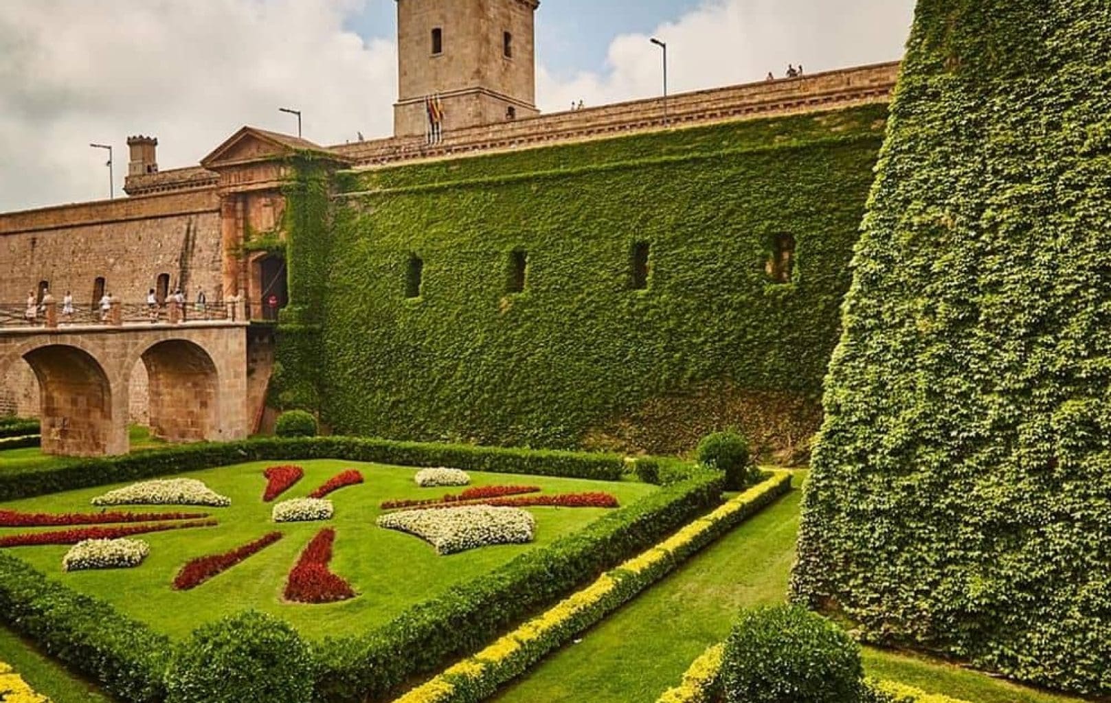 Montjuïc Castle, a historic stone fortress in Barcelona, features a tower and an arched bridge surrounded by manicured gardens and ivy-clad walls, making it a must-see tourist attraction.