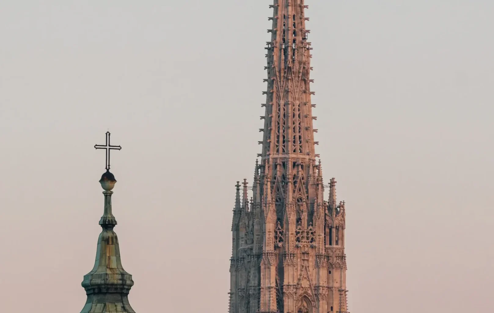 A church steeple with a cross and the tall, ornate spire of St. Stephens Cathedral in Vienna rise against a pale sky at sunset.