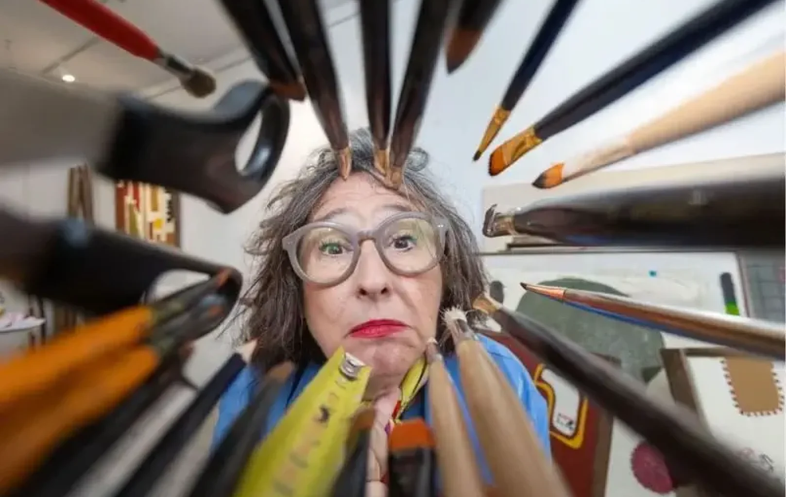 A woman with glasses looks through a circular arrangement of pencils and paintbrushes in an art studio.